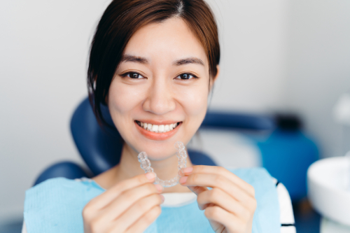 Smiling woman sits in dental surgery holding a clear aligner just below her chin
