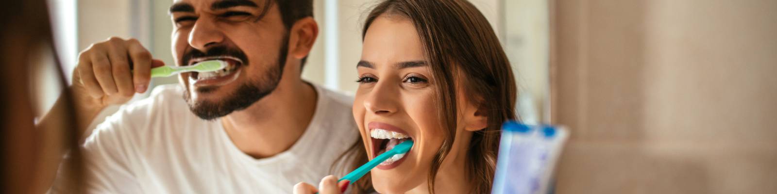 A man and a woman stand side by side brushing their teeth together. The image shows their reflection in the mirror over the sink.